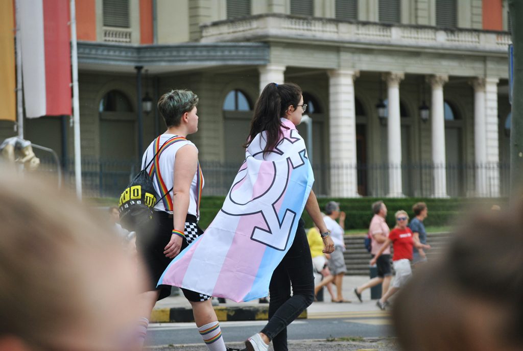 Two people walking down a busy street, one has a trans rights flag wrapped around their shoulders